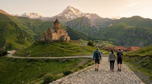 Trois randonneurs sur un chemin pavé vers l'église de Gergeti, avec le mont Kazbek enneigé en arrière-plan sous un ciel clair.