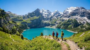Trois randonneurs avec des sacs à dos contemplent le lac turquoise d'Oeschinen entouré de montagnes enneigées.