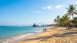 Couple marchant sur une plage de sable fin à Bali avec le temple Pura Gede Pemuteran et un volcan en arrière-plan.